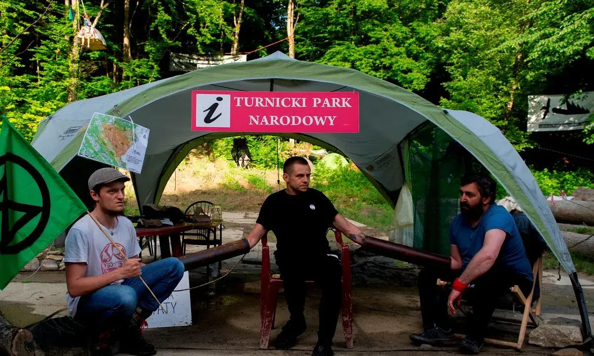 Trzech mężczyzn siedzi pod namiotem z napisem "Turnicki Park Narodowy". Obok zielona flaga z symbolem.