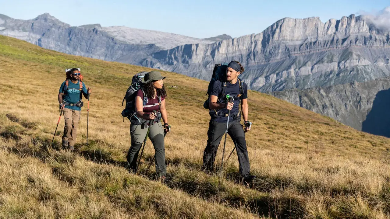 Trzy osoby z plecakami i kijkami trekkingowymi idące po trawiastym zboczu górskim.
