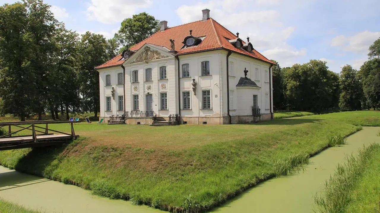 Pałac w Choroszczy otoczony fosą i zielenią, z drewnianym mostkiem prowadzącym do wejścia.