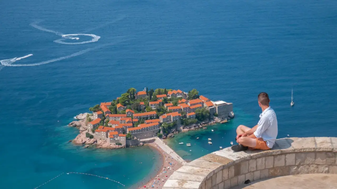 Czarnogóra na co uważać? Widok na Sveti Stefan, turkusowe morze i plażę. Mężczyzna siedzi na murku, kontemplując krajobraz.