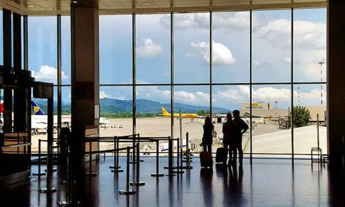 Widok z okna na **bergamo lotnisko**. Trzy osoby z walizkami czekają na samolot. W tle żółty samolot DHL i góry.