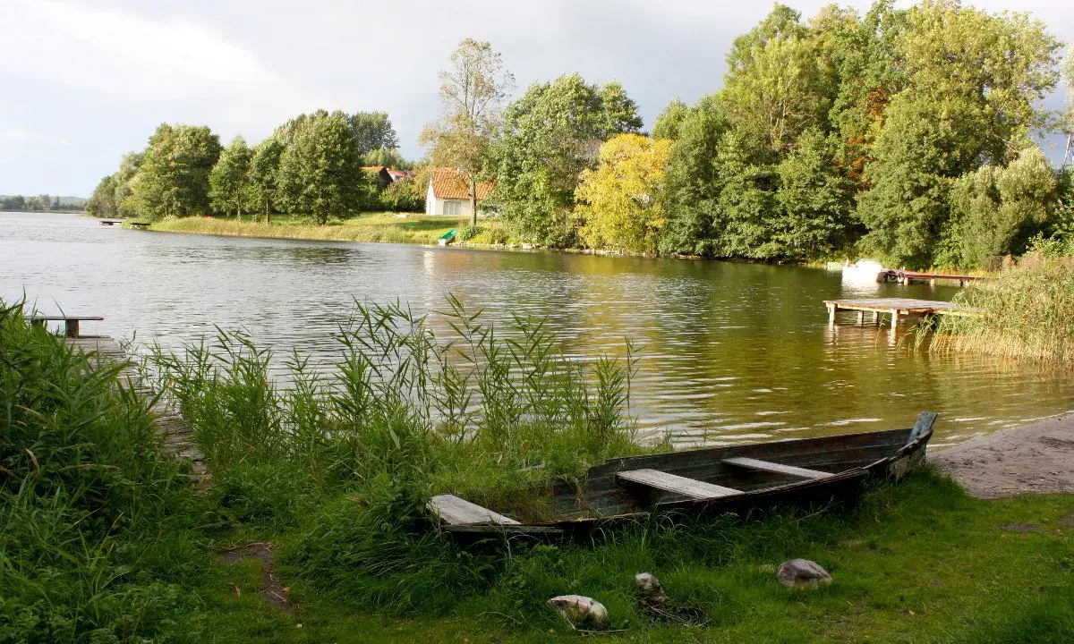 Łódka na brzegu, w tle zielone drzewa i domki nad wodą. Widok na największe jezioro w Polsce.