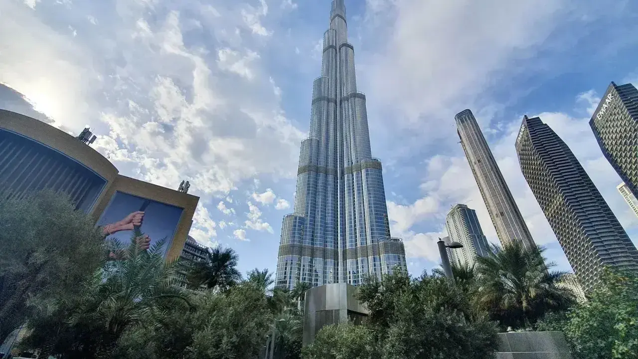 Burj Khalifa, najwyższy budynek w Dubaju, wznosi się dumnie na tle błękitnego nieba z chmurami. Otoczony zielenią i innymi drapaczami chmur.