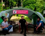 Trzech mężczyzn siedzi pod namiotem z napisem "Turnicki Park Narodowy". Obok zielona flaga z symbolem.