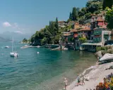 Urokliwe como miasto nad jeziorem, z kolorowymi domami na wzgórzu, łodziami na wodzie i dziećmi bawiącymi się na plaży.