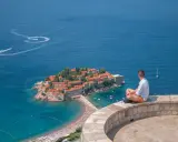 Czarnogóra na co uważać? Widok na Sveti Stefan, turkusowe morze i plażę. Mężczyzna siedzi na murku, kontemplując krajobraz.