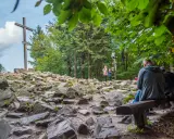 Kamienista ścieżka prowadzi do wejścia na Łysicę, gdzie stoi drewniany krzyż. Ludzie odpoczywają na ławce, podziwiając widok.