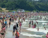 Tłumy ludzi na plaży i w morzu, szukają idealnego miejsca gdzie na wakacje w Polsce. W tle zjeżdżalnie wodne i las.