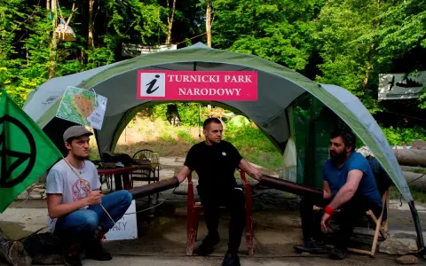 Trzech mężczyzn siedzi pod namiotem z napisem "Turnicki Park Narodowy". Obok zielona flaga z symbolem.