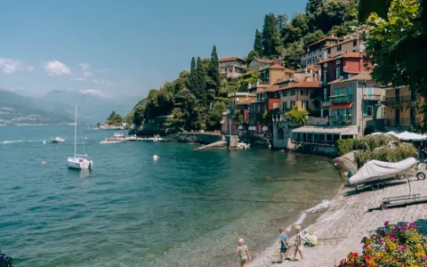 Urokliwe como miasto nad jeziorem, z kolorowymi domami na wzgórzu, łodziami na wodzie i dziećmi bawiącymi się na plaży.