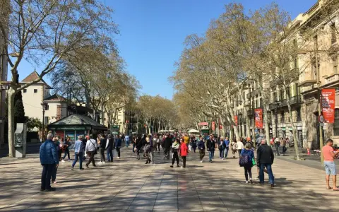 Tłum ludzi spaceruje po słynnej alei La Rambla w Barcelonie, otoczonej drzewami i zabytkowymi budynkami.