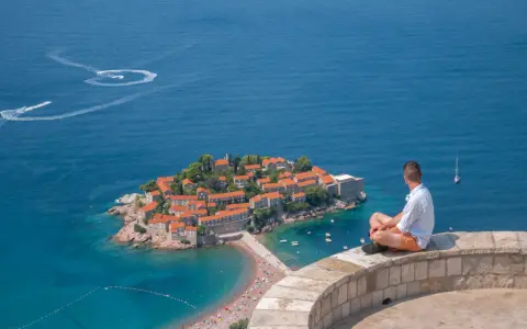 Czarnogóra na co uważać? Widok na Sveti Stefan, turkusowe morze i plażę. Mężczyzna siedzi na murku, kontemplując krajobraz.