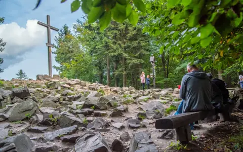 Kamienista ścieżka prowadzi do wejścia na Łysicę, gdzie stoi drewniany krzyż. Ludzie odpoczywają na ławce, podziwiając widok.