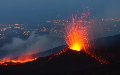 Spektakularna erupcja Etny nocą, z płynącą lawą i deszczem iskier nad oświetlonym miastem.