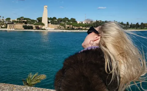 Kobieta z blond włosami podziwia pomnik w Brindisi. W tle błękitne morze i błękitne niebo.