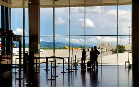Widok z okna na **bergamo lotnisko**. Trzy osoby z walizkami czekają na samolot. W tle żółty samolot DHL i góry.