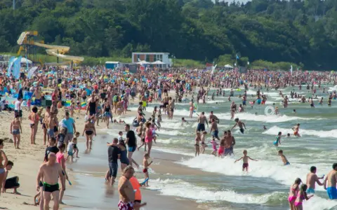 Tłumy ludzi na plaży i w morzu, szukają idealnego miejsca gdzie na wakacje w Polsce. W tle zjeżdżalnie wodne i las.