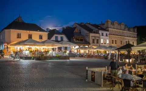 Wieczorny rynek w Kazimierzu Dolnym tętni życiem. Ludzie siedzą przy stolikach pod parasolami, ciesząc się atmosferą.