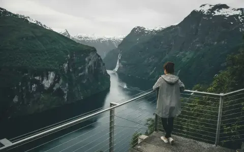 Kobieta podziwia majestatyczny **fjord** z wodospadem i ośnieżonymi szczytami w tle.
