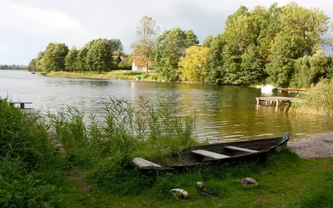 Łódka na brzegu, w tle zielone drzewa i domki nad wodą. Widok na największe jezioro w Polsce.