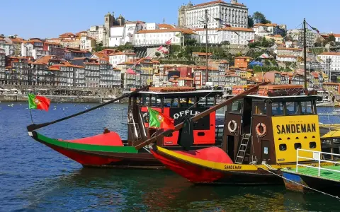 Tradycyjne łodzie Rabelo cumują w malowniczym porcie miasta Porto, z kolorowymi domami na wzgórzu w tle.