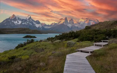 Drewniana ścieżka prowadzi przez zielone wzgórza ku turkusowemu jezioru i ośnieżonym górom. Zachód słońca maluje niebo nad **Tierra del Fuego** odcieniami różu i pomarańczy.