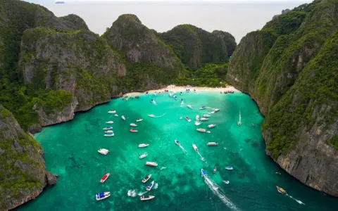 Maya Bay tętni życiem, pełna łodzi i turystów na piaszczystej plaży, otoczona stromymi, porośniętymi zielenią klifami.