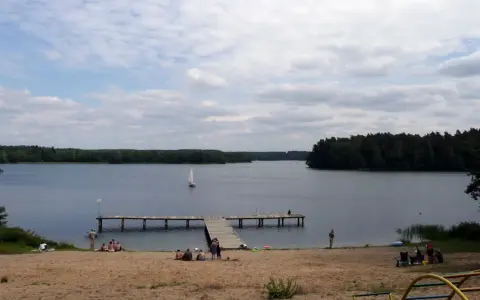 Spokojny dzień na plaży nad jeziorem, żaglówka na wodzie, drewniane molo. Pojezierze Brodnickie zaprasza do wypoczynku.