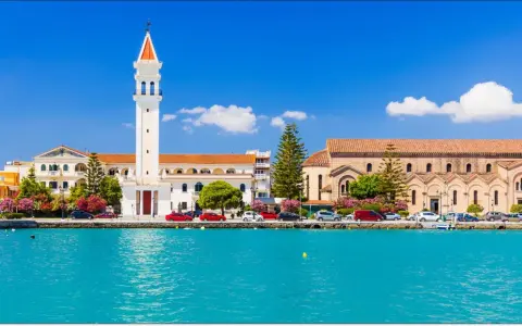 Widok na miasto Zakynthos z turkusową wodą i budynkami, w tym kościołem z charakterystyczną wieżą.