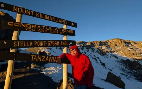 Mężczyzna w czerwonej kurtce przy znaku "Mount Kilimanjaro", najwyższy szczyt Afryki. Gratulacje, jesteś na Stella Point 5756 m n.p.m.