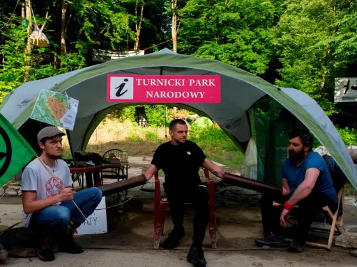Trzech mężczyzn siedzi pod namiotem z napisem "Turnicki Park Narodowy". Obok zielona flaga z symbolem.