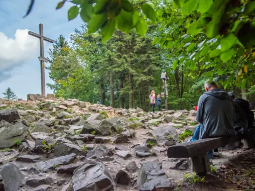Kamienista ścieżka prowadzi do wejścia na Łysicę, gdzie stoi drewniany krzyż. Ludzie odpoczywają na ławce, podziwiając widok.