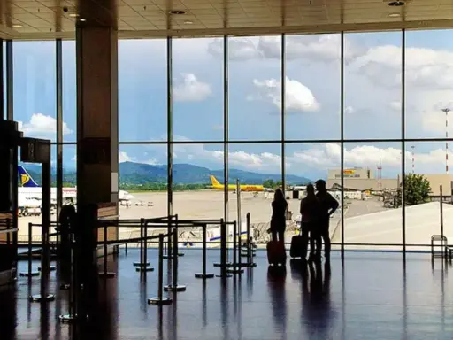 Widok z okna na **bergamo lotnisko**. Trzy osoby z walizkami czekają na samolot. W tle żółty samolot DHL i góry.
