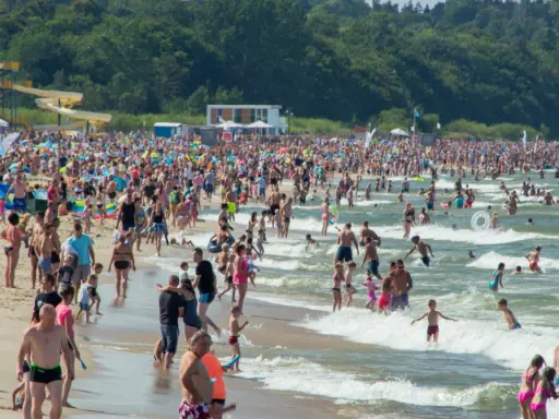 Tłumy ludzi na plaży i w morzu, szukają idealnego miejsca gdzie na wakacje w Polsce. W tle zjeżdżalnie wodne i las.