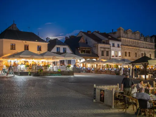 Wieczorny rynek w Kazimierzu Dolnym tętni życiem. Ludzie siedzą przy stolikach pod parasolami, ciesząc się atmosferą.