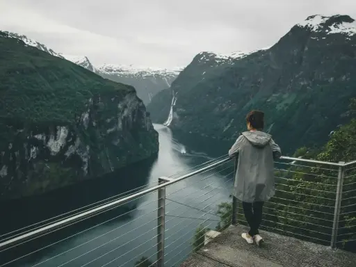 Kobieta podziwia majestatyczny **fjord** z wodospadem i ośnieżonymi szczytami w tle.