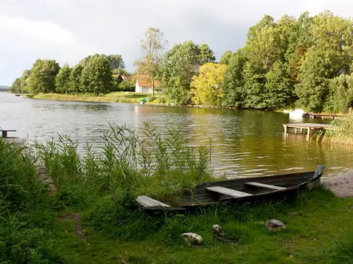 Łódka na brzegu, w tle zielone drzewa i domki nad wodą. Widok na największe jezioro w Polsce.