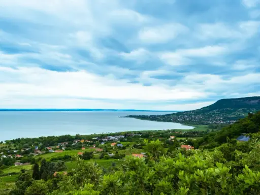 Widok na Balaton z góry, z zielonymi wzgórzami i domkami. W oddali widać jezioro i plażę.