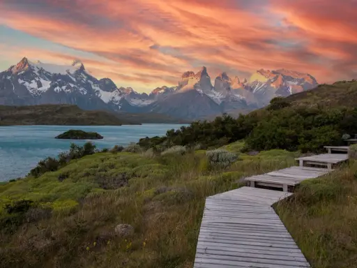 Drewniana ścieżka prowadzi przez zielone wzgórza ku turkusowemu jezioru i ośnieżonym górom. Zachód słońca maluje niebo nad **Tierra del Fuego** odcieniami różu i pomarańczy.