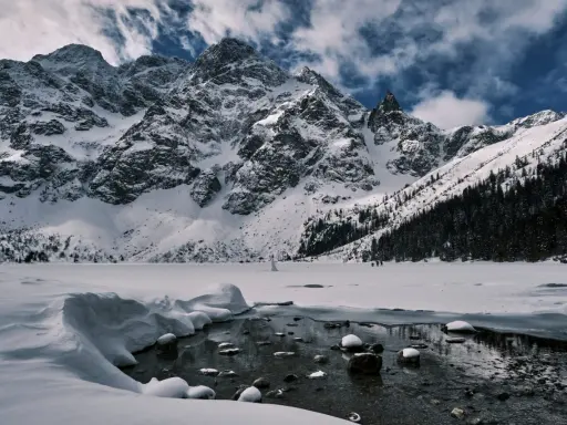 Morskie Oko zimową porą. Lodowate wody jeziora odbijają ośnieżone szczyty Tatr, a na horyzoncie widać ludzi spacerujących po zamarzniętej tafli.
