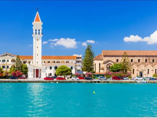 Widok na miasto Zakynthos z turkusową wodą i budynkami, w tym kościołem z charakterystyczną wieżą.
