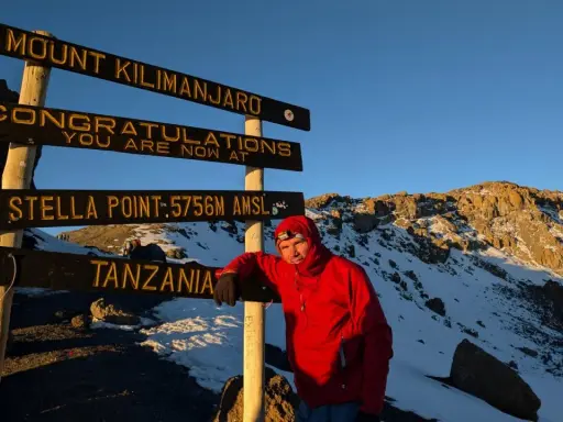 Mężczyzna w czerwonej kurtce przy znaku "Mount Kilimanjaro", najwyższy szczyt Afryki. Gratulacje, jesteś na Stella Point 5756 m n.p.m.