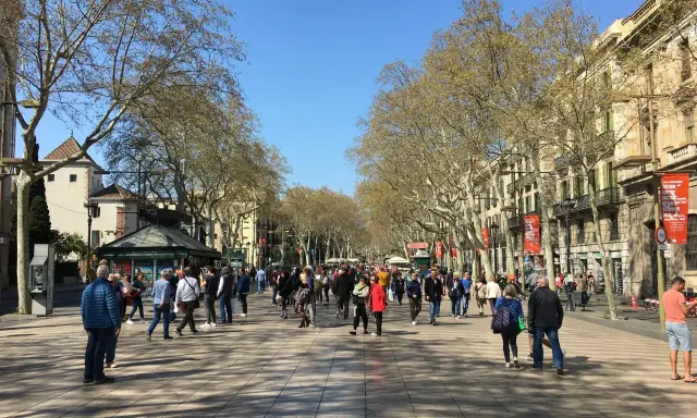Tłum ludzi spaceruje po słynnej alei La Rambla w Barcelonie, otoczonej drzewami i zabytkowymi budynkami.