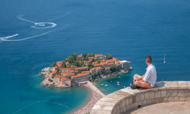 Czarnogóra na co uważać? Widok na Sveti Stefan, turkusowe morze i plażę. Mężczyzna siedzi na murku, kontemplując krajobraz.
