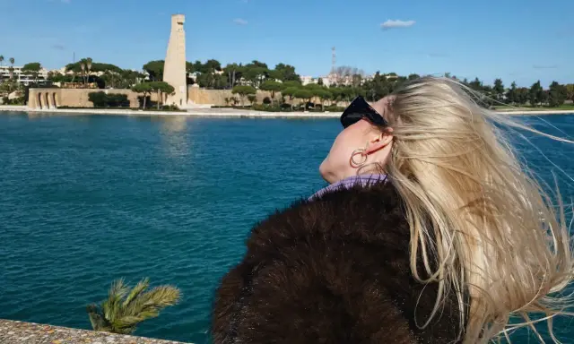 Kobieta z blond włosami podziwia pomnik w Brindisi. W tle błękitne morze i błękitne niebo.