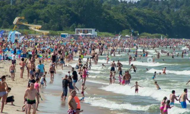 Tłumy ludzi na plaży i w morzu, szukają idealnego miejsca gdzie na wakacje w Polsce. W tle zjeżdżalnie wodne i las.