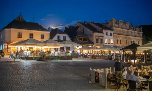 Wieczorny rynek w Kazimierzu Dolnym tętni życiem. Ludzie siedzą przy stolikach pod parasolami, ciesząc się atmosferą.