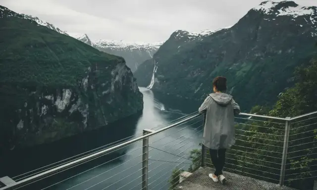 Kobieta podziwia majestatyczny **fjord** z wodospadem i ośnieżonymi szczytami w tle.