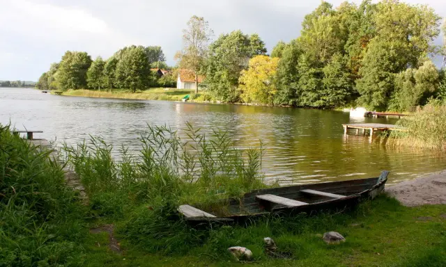 Łódka na brzegu, w tle zielone drzewa i domki nad wodą. Widok na największe jezioro w Polsce.
