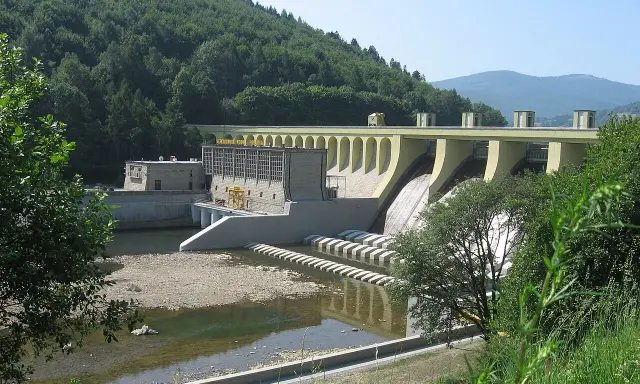 Zapora wodna w Porąbce, z charakterystycznymi łukami i budynkiem elektrowni. Otoczona zielonymi wzgórzami.